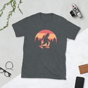 May include: A dark heather grey t-shirt with a retro-style graphic of Bigfoot roller skating in front of a sunset. The graphic is in shades of red, orange, and yellow.