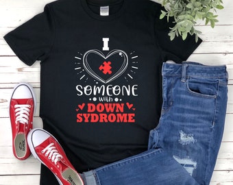 Op de afbeelding: Zwart T-shirt met een witte afbeelding van een hart met een puzzelstukje erin. De tekst luidt "Ik hou van iemand met het syndroom van Down".