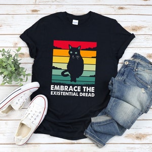 May include: Black t-shirt with a retro-style graphic of a black cat sitting in front of a sunset. The text "Embrace the Existential Dread" is printed below the cat.