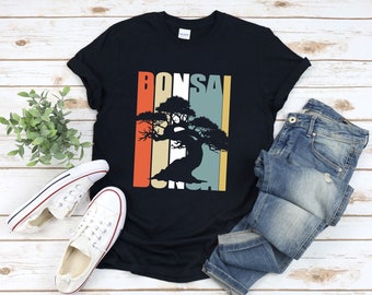 Könnte beinhalten: Ein schwarzes T-Shirt mit einem Vintage-Grafikdruck eines Bonsai-Baums. Die Grafik zeigt eine Silhouette des Baumes vor einem bunten gestreiften Hintergrund. Der Text "Bonsai" ist vertikal auf dem Shirt gedruckt.