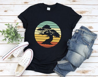 Könnte beinhalten: Ein schwarzes T-Shirt mit einer Retro-Grafik eines Bonsai-Baums in einem Sonnenuntergang. Die Grafik ist in Orange-, Gelb- und Grüntönen gehalten.