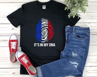 Puede incluir: Camiseta negra con un diseño de huella dactilar blanca con los colores de la bandera francesa. El texto "It's in my DNA" está impreso debajo del diseño.