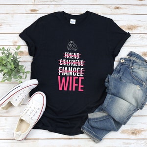 Puede incluir: Camiseta negra con texto blanco que dice "Friend, Girlfriend, Fiancee, Wife" con una l&iacute;nea rosa que atraviesa las tres primeras palabras y un anillo rosa sobre la palabra "Friend". La palabra "Wife" est&aacute; en rosa.