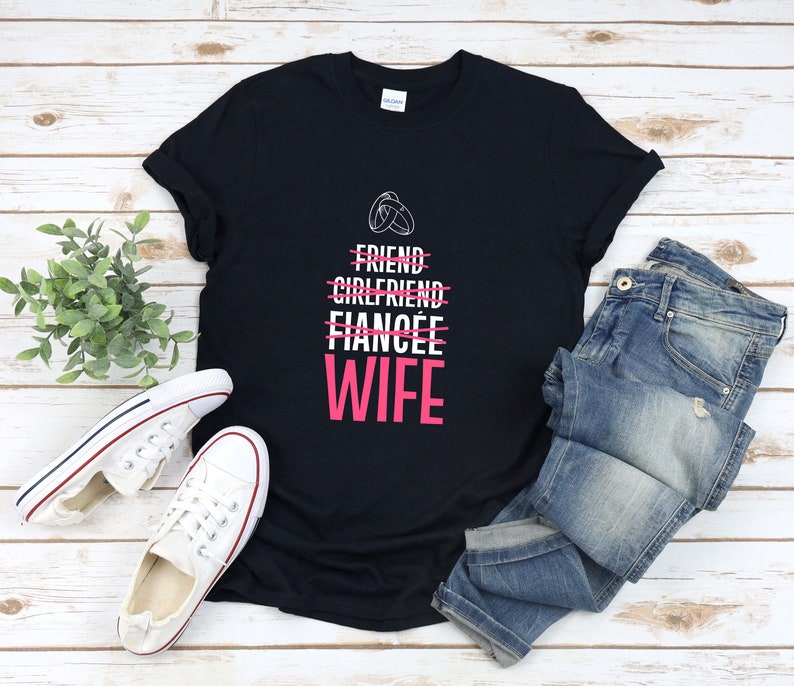 Puede incluir: Camiseta negra con texto blanco que dice "Friend, Girlfriend, Fiancee, Wife" con una l&iacute;nea rosa que atraviesa las tres primeras palabras y un anillo rosa sobre la palabra "Friend". La palabra "Wife" est&aacute; en rosa.