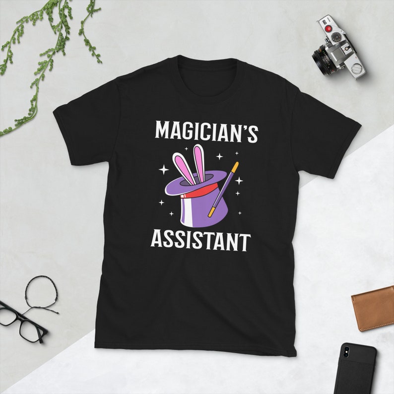 May include: Black t-shirt with a white graphic of a magician's hat with a rabbit sticking out of it and a magic wand. The text "MAGICIAN'S ASSISTANT" is printed below the graphic.