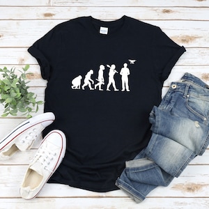 Może przedstawiać: Czarny T-shirt z białym nadrukiem przedstawiającym ewolucję człowieka, kończącą się na człowieku trzymającym drona. Grafika jest humorystycznym spojrzeniem na ewolucję człowieka, pokazującym, jak drony stały się ważną częścią współczesnego życia.