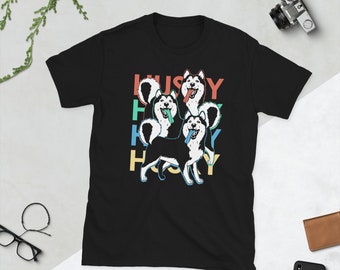 Op de afbeelding: Zwart T-shirt met een afbeelding van drie husky-honden met hun tongen uit, omringd door het woord "Husky" in regenboogkleuren.