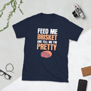 May include: Navy blue t-shirt with white text that reads "Feed me brisket and tell me I'm pretty". A cartoon image of a slice of brisket is below the text.