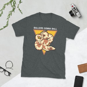 Op de afbeelding: Een donkergrijs T-shirt met een witte driehoek en de tekst "Ballers Gonna Ball" in wit. Binnen de driehoek is een cartoon afbeelding van een bruine en witte slang.