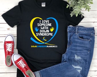 Op de afbeelding: Zwart T-shirt met een geel en blauw hartontwerp. Het hart heeft de tekst "Ik hou van iemand met het syndroom van Down" en twee handafdrukken. Onder het hart staat de tekst "Down-syndroom bewustzijn".