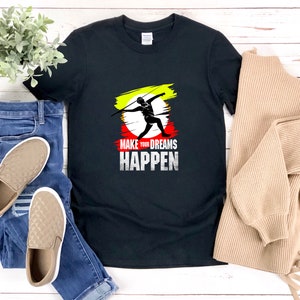 May include: Black t-shirt with a graphic of a person throwing a javelin. The graphic is surrounded by a yellow, red, and white paint splatter. The text "Make your dreams happen" is printed below the graphic.