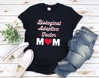 May include: Black t-shirt with white text that says "Biological, Adoptive, Foster" with lines through each word. The text "MOM" is below the crossed out words with a red heart between the M's.