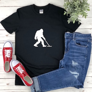 Puede incluir: Camiseta negra con una silueta blanca de Bigfoot sosteniendo un detector de metales. La camiseta está sobre una superficie de madera blanca con un par de zapatillas rojas y unos vaqueros azules.