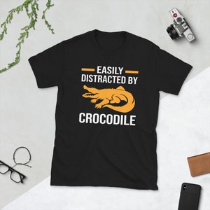 Op de afbeelding: Zwart T-shirt met de tekst "EASILY DISTRACTED BY CROCODILE" in het wit. Het shirt heeft een oranje krokodilafbeelding. Een camera, bril en portemonnee zijn zichtbaar op de achtergrond.