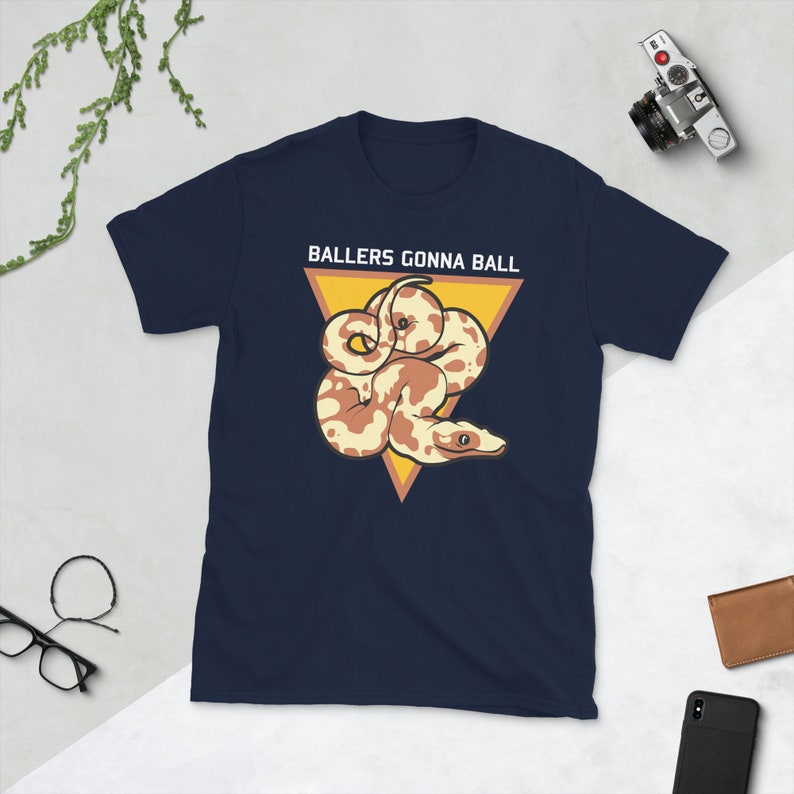 Op de afbeelding: Een marineblauw t-shirt met een witte driehoek en de tekst "Ballers Gonna Ball" in wit. Binnen de driehoek is een cartoon afbeelding van een bruine en witte slang.