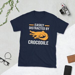 Op de afbeelding: Marineblauw T-shirt met de tekst "EASILY DISTRACTED BY CROCODILE" in het wit en een oranje krokodilafbeelding. Het shirt is gemaakt van een zacht materiaal.