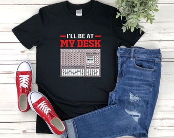 May include: Black t-shirt with the text "I'll be at my desk" in white and a graphic of a music production mixer in pink. The shirt is being modeled on a person wearing blue jeans.