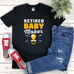 Può includere: Maglietta nera con testo bianco che recita "Retired Baby Maker" e un'immagine di ciuccio giallo.