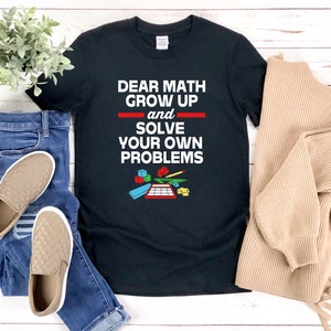 May include: Black t-shirt with the text "Dear Math Grow Up and Solve Your Own Problems" in white letters. The text is underlined in red. There are colorful toy blocks and a pencil on a calculator below the text.