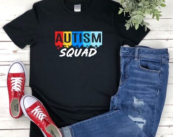 May include: Black t-shirt with the words "AUTISM SQUAD" in colorful puzzle piece design. The shirt is paired with blue jeans and red canvas sneakers with white laces. A small green plant is in the background.