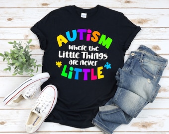 Puede incluir: Camiseta negra con el texto "AUTISMO donde las pequeñas cosas nunca son PEQUEÑAS" en colores arcoíris. El texto está rodeado de símbolos de piezas de rompecabezas.