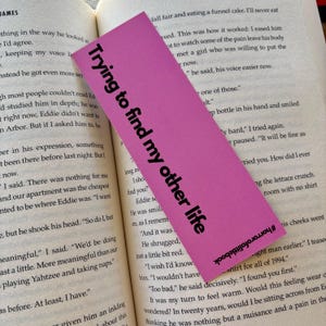May include: A pink bookmark with the text "Trying to find my other life" in black lettering. The bookmark is slightly curled at the bottom and has a hashtag at the bottom right corner.