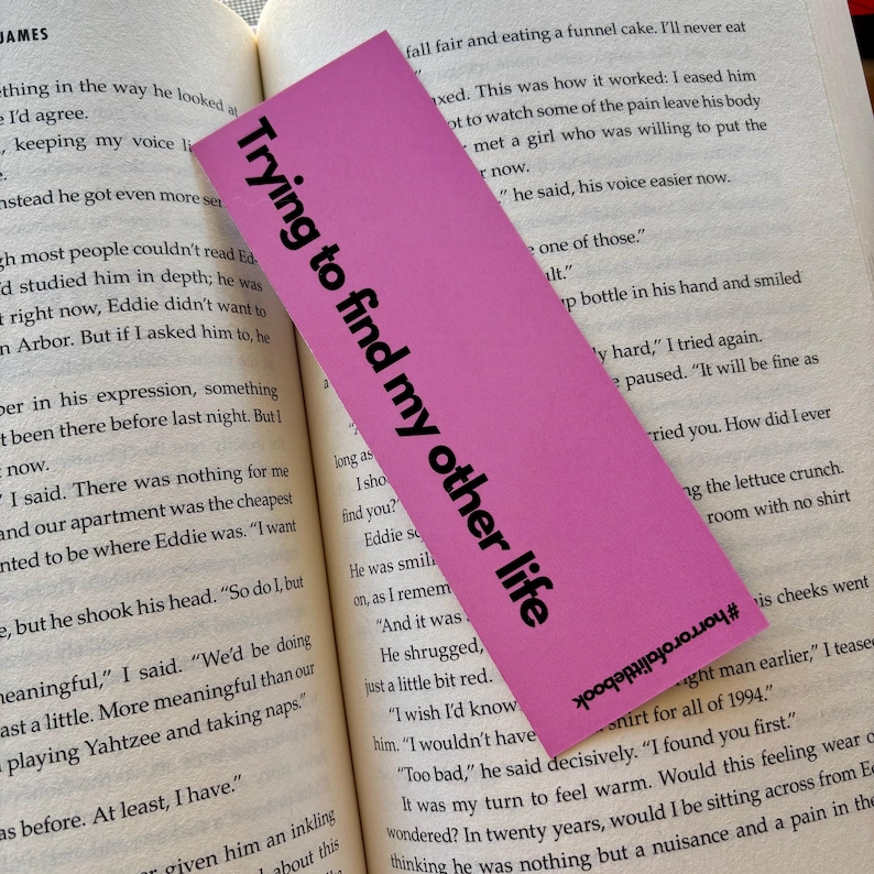 May include: A pink bookmark with the text "Trying to find my other life" in black lettering. The bookmark is slightly curled at the bottom and has a hashtag at the bottom right corner.