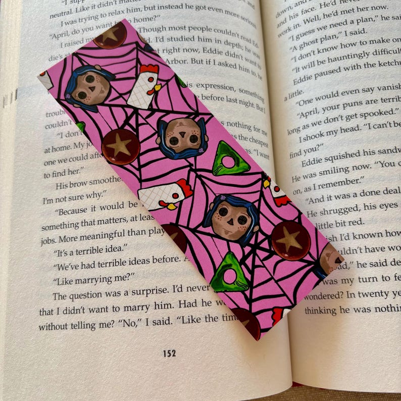 May include: Pink bookmark with a repeating pattern of a girl with black hair, a spiderweb, and a green triangle. The girl is wearing a blue dress and has a red bow on her hair. The bookmark also features a red star and a white chicken.