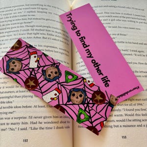 May include: Pink bookmark with a repeating pattern of a spiderweb, a doll with black hair, a chicken, and a star. The text "Trying to find my other life" is printed on the bookmark.