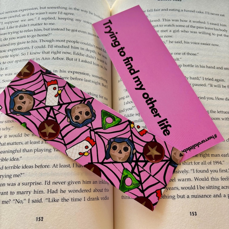 May include: Pink bookmark with a repeating pattern of a spiderweb, a doll with black hair, a chicken, and a star. The text "Trying to find my other life" is printed on the bookmark.