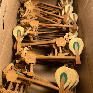 May include: A collection of wooden piano hammers, showing the felt heads and wooden levers. The hammers are arranged inside a cardboard box, with the wooden components in various positions. The felt heads have a green stripe.