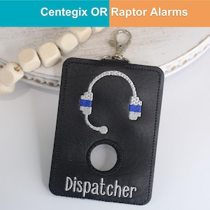 May include: A black rectangular leather-like holder with a silver metal swivel clasp. It features an embroidered silver and blue headset icon above a circular cutout and the word Dispatcher embroidered in silver thread at the bottom edge.