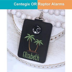 May include: Black rectangular key fob with a silver metal clasp. The fob features a silver button and an embroidered design of two palm trees and the name "Elizabeth" in green. The text "Centegix OR Raptor Alarms" is at the top.