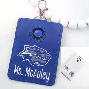 May include: Blue leather key chain with a wolf mascot and the name "Ms. McAuley" embroidered in white. The keychain has a blue button and a silver clip. A white square device with a QR code and the text "CENTEGYX" is also visible.
