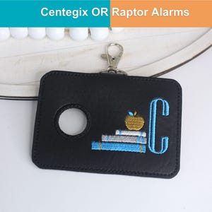 May include: A black rectangular holder for Centegix OR Raptor Alarms featuring a circular cutout, a silver metal lobster clasp, and embroidery of a stack of books topped with a gold apple next to a large light blue letter C.