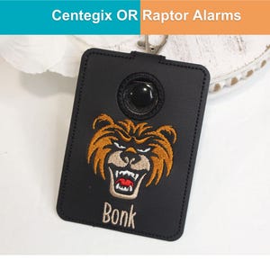 May include: A black rectangular holder for Centegix OR Raptor Alarms featuring an embroidered brown bear face with an open mouth showing teeth and the word Bonk stitched in white thread below the bear. It includes a metal clip at the top.