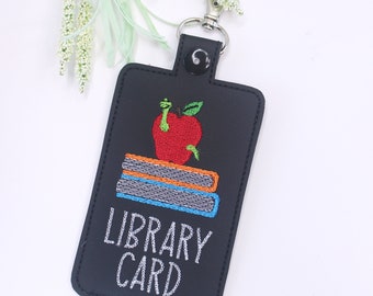 May include: Black leather library card holder with a red apple, green worm, and stacked books embroidered on it. The text "LIBRARY CARD" is embroidered in white below the image.