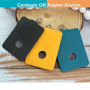 May include: Three rectangular protective cases for Centegix OR Raptor Alarms, each featuring a circular cutout and a metal swivel clasp. The cases are available in black, yellow, and teal, with visible stitching along the edges of each holder.