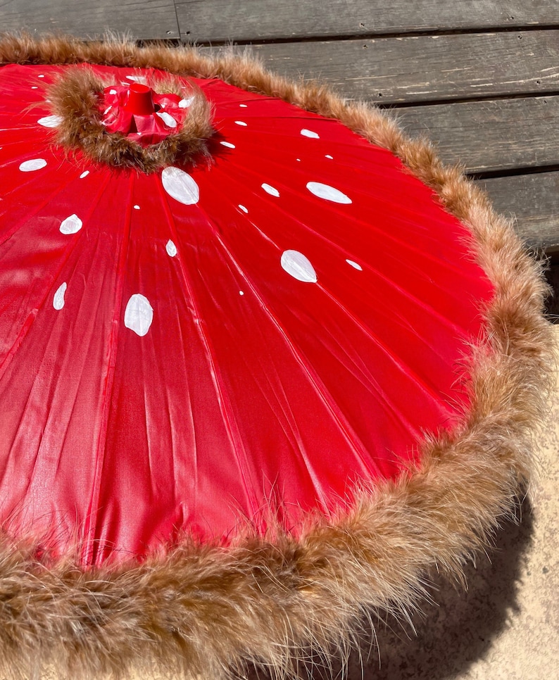 Pode incluir: Um guarda-sol vermelho com bolinhas brancas, assemelhando-se a um cogumelo. A borda &eacute; adornada com pele marrom. O guarda-sol est&aacute; aberto, mostrando a parte inferior.