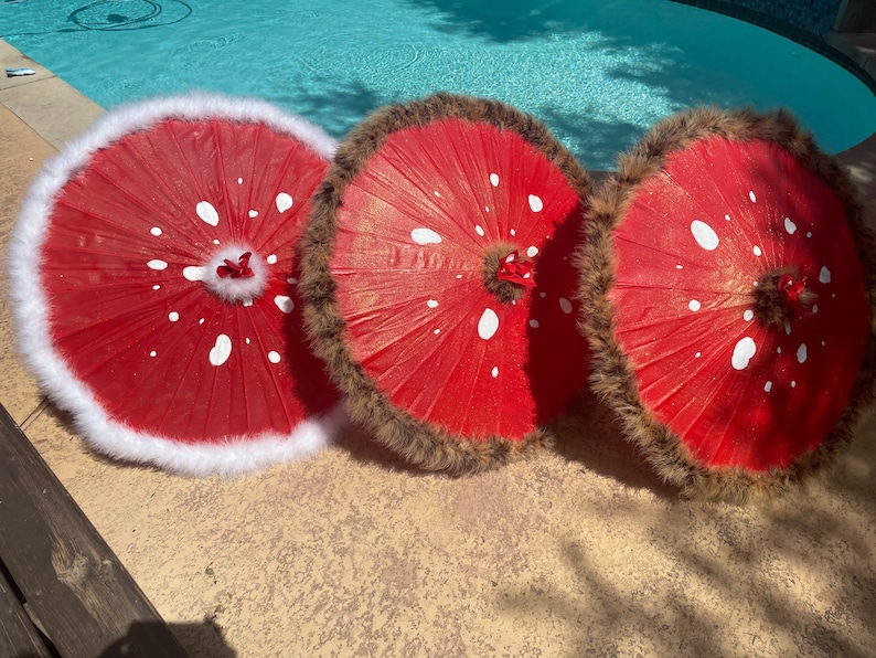Pode incluir: Tr&ecirc;s guarda-s&oacute;is vermelhos com bolinhas brancas e acabamentos decorativos. Um guarda-sol tem acabamento em pele sint&eacute;tica branca, enquanto os outros dois t&ecirc;m acabamento em pele sint&eacute;tica castanha. Os guarda-s&oacute;is est&atilde;o dispostos sobre uma superf&iacute;cie de pedra.