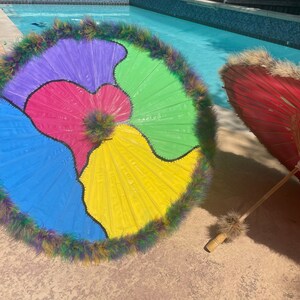 Pode incluir: Um guarda-sol colorido com um design segmentado em azul, roxo, rosa, verde e amarelo, adornado com uma borda de penas fofas. Um segundo guarda-sol vermelho com cabo de bambu tamb&eacute;m &eacute; vis&iacute;vel.
