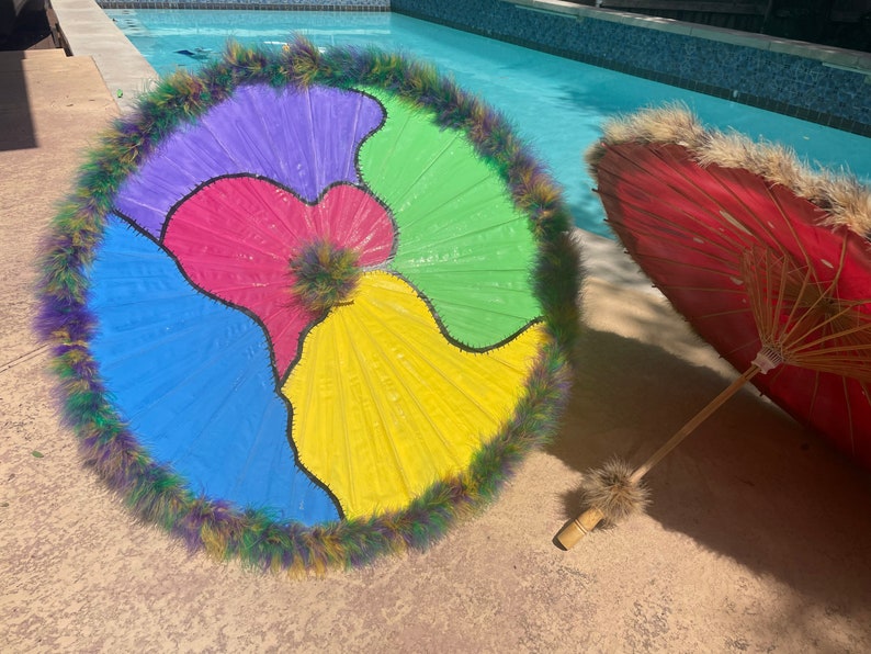Pode incluir: Um guarda-sol colorido com um design segmentado em azul, roxo, rosa, verde e amarelo, adornado com uma borda de penas fofas. Um segundo guarda-sol vermelho com cabo de bambu tamb&eacute;m &eacute; vis&iacute;vel.