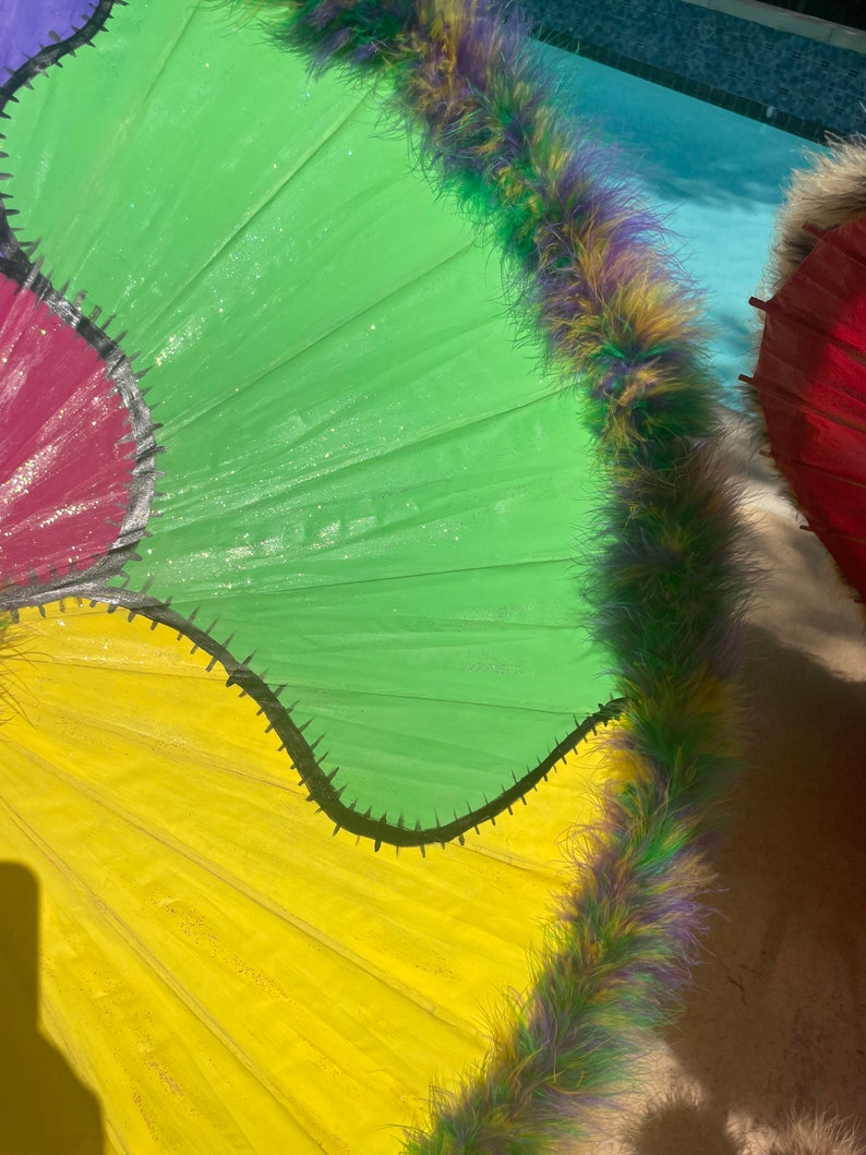 Pode incluir: Close-up de guarda-s&oacute;is coloridos com um design vibrante. Os guarda-s&oacute;is apresentam pain&eacute;is verdes brilhantes, amarelos e vermelhos, delineados com um padr&atilde;o de zigue-zague preto. Cada guarda-sol &eacute; adornado com penas fofas multicoloridas.