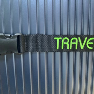 May include: Close-up of a navy blue suitcase with vertical ribbed detailing. A black strap with a buckle is secured around the suitcase. The word "TRAVEL" is embroidered in bright green on the strap.
