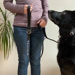 May include: Black dog leash with the word "TROUBLE" printed in white. The leash is held by a person wearing blue jeans and a purple long-sleeve shirt. A black dog is looking up at the person.