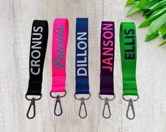 May include: Five fabric wristlet keychains with metal lobster clasps. The black strap reads CRONUS, the neon pink strap reads Patricia, the blue strap reads DILLON, the purple strap reads JANSON, and the green strap reads ELLIS.
