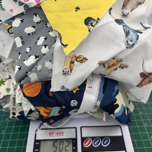 May include: Assorted fabric remnants featuring animal and space designs, such as cows, sheep, and dinosaurs. The fabrics are placed on a digital scale, displaying a weight of 502. The scale has buttons for power, mode, and tare.