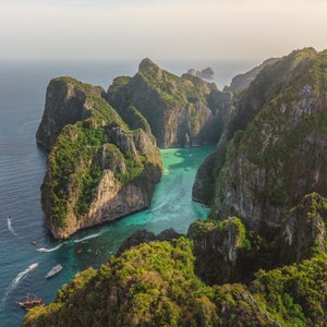 May include: An aerial view of a tropical island with lush green vegetation and towering limestone cliffs. The turquoise waters of the ocean surround the island, and several boats are visible in the water.