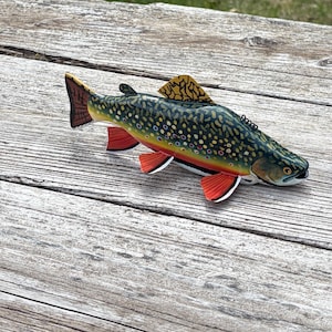 May include: A wooden fish figurine with a detailed painted design. The fish has a dark green back with yellow and brown spots, red fins, and a yellow dorsal fin. It rests on a weathered wooden surface.