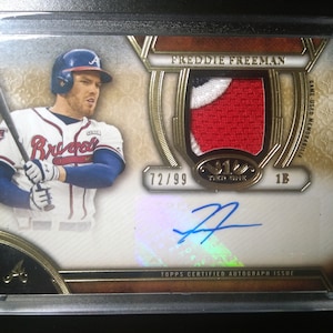 May include: A baseball card featuring Freddie Freeman of the Atlanta Braves. The card is numbered 72/99 and includes a game-used jersey patch. The card is signed by Freddie Freeman.